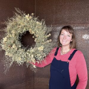 Carlyn holding a cream and sage dried wreath against a rusted steel wall.