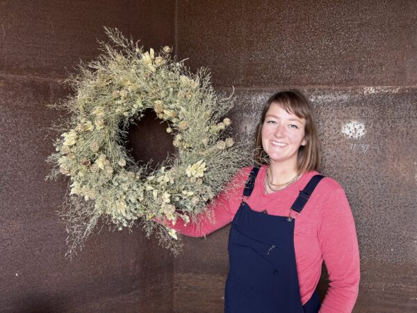 Carlyn holding a cream and sage dried wreath against a rusted steel wall.