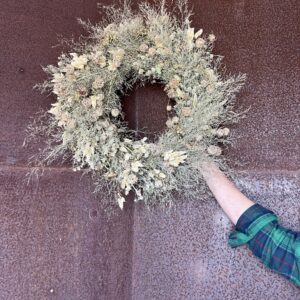 A hand holding a small cream dried wreath against a rusted steel door.