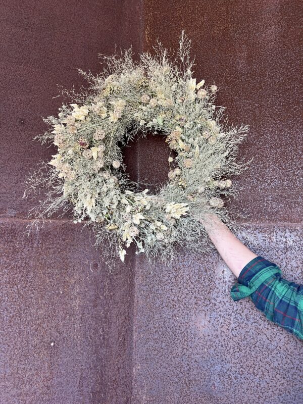 A hand holding a small cream dried wreath against a rusted steel door.
