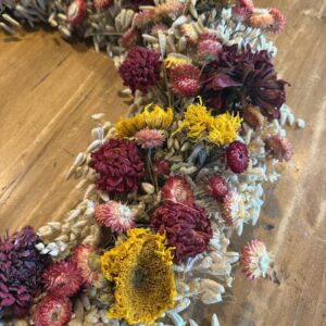 A dried wreath in strawflower, gomphrena, statice and a sunflower head, laid flat on a wood table.