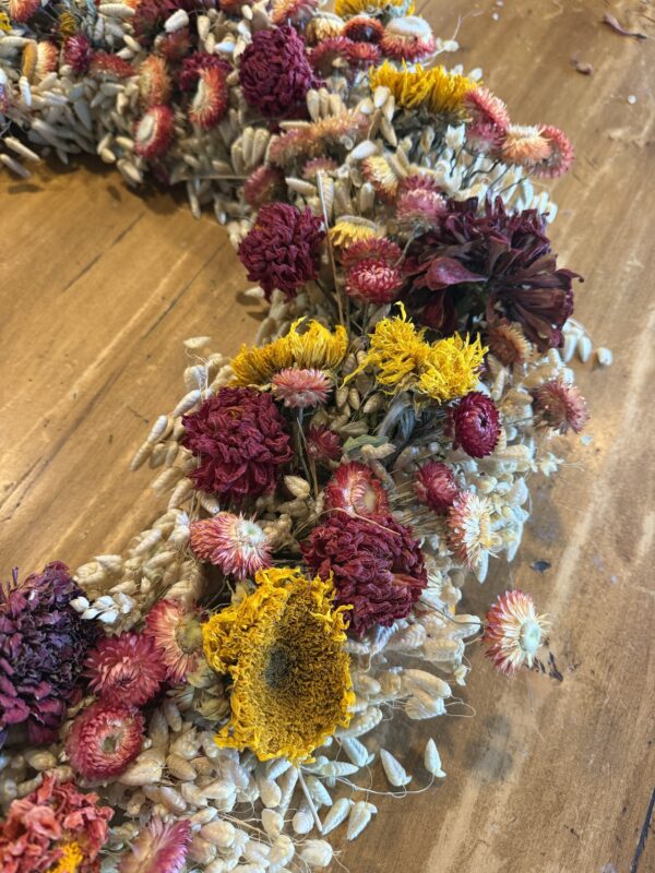 A dried wreath in strawflower, gomphrena, statice and a sunflower head, laid flat on a wood table.